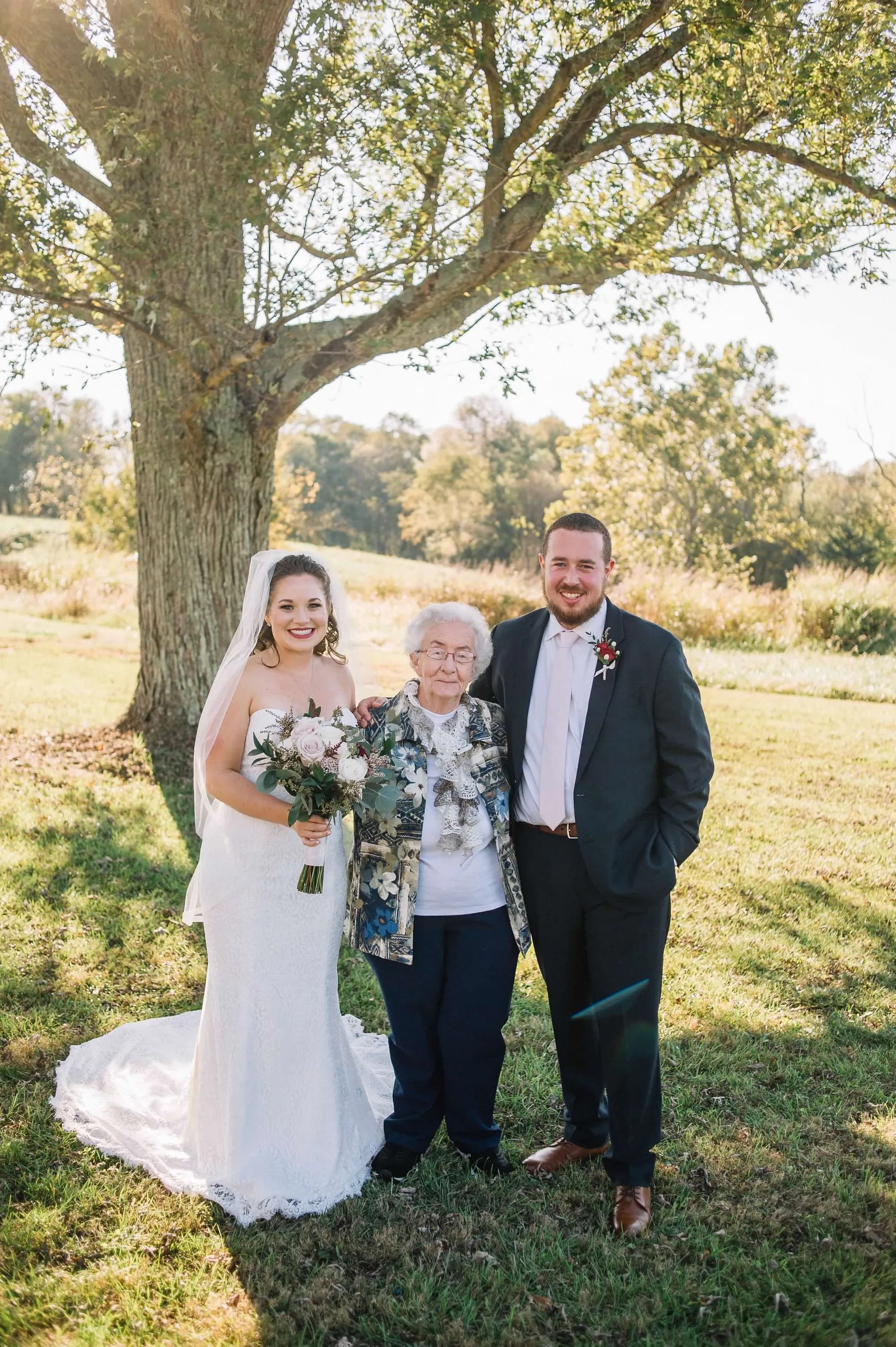 Bride and groom pose with elderly grandmother under a large tree on sunlit estate grounds