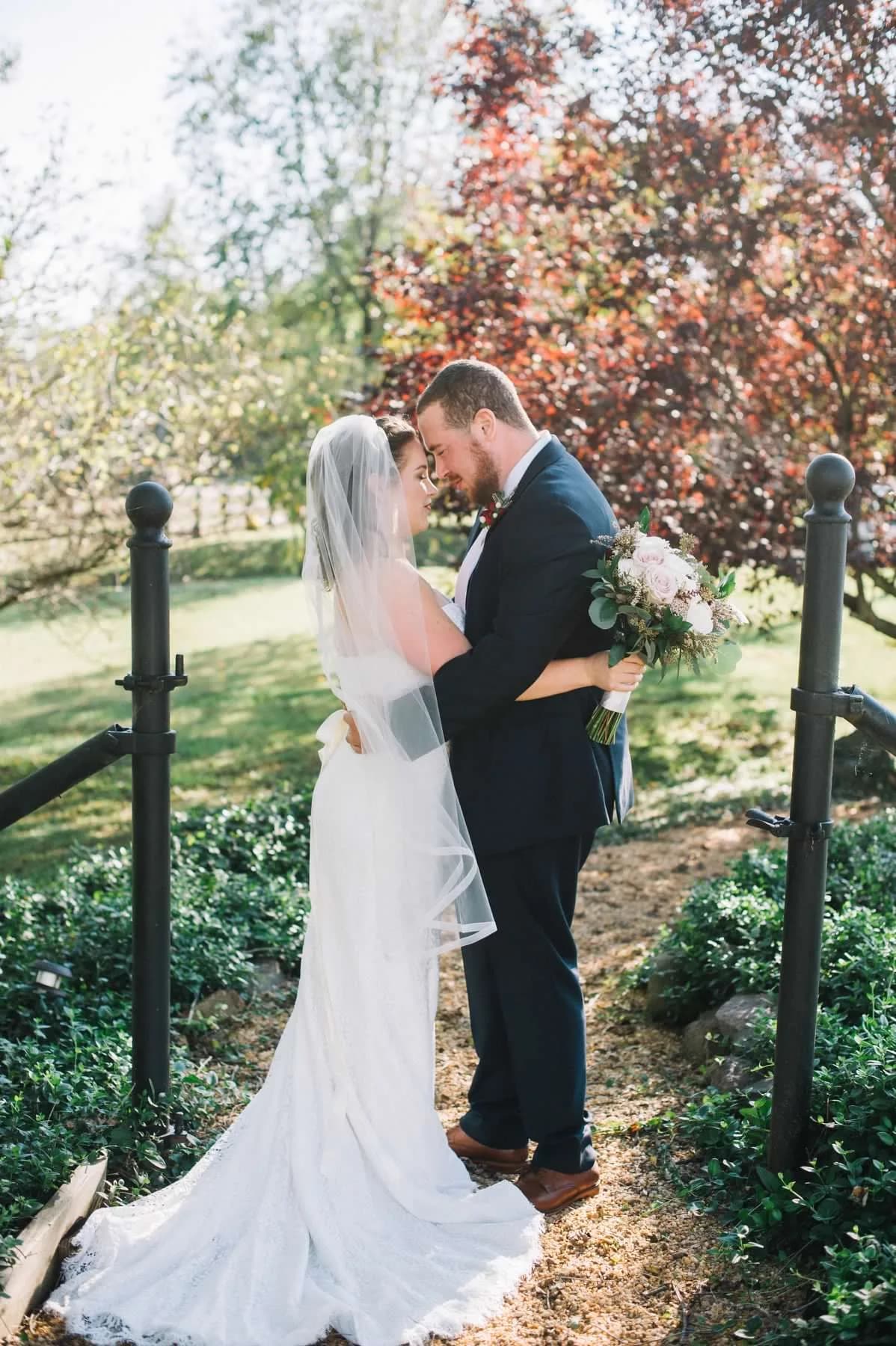 Bride and groom share an intimate moment on garden path at Rixey Manor, surrounded by lush greenery and red-leafed trees