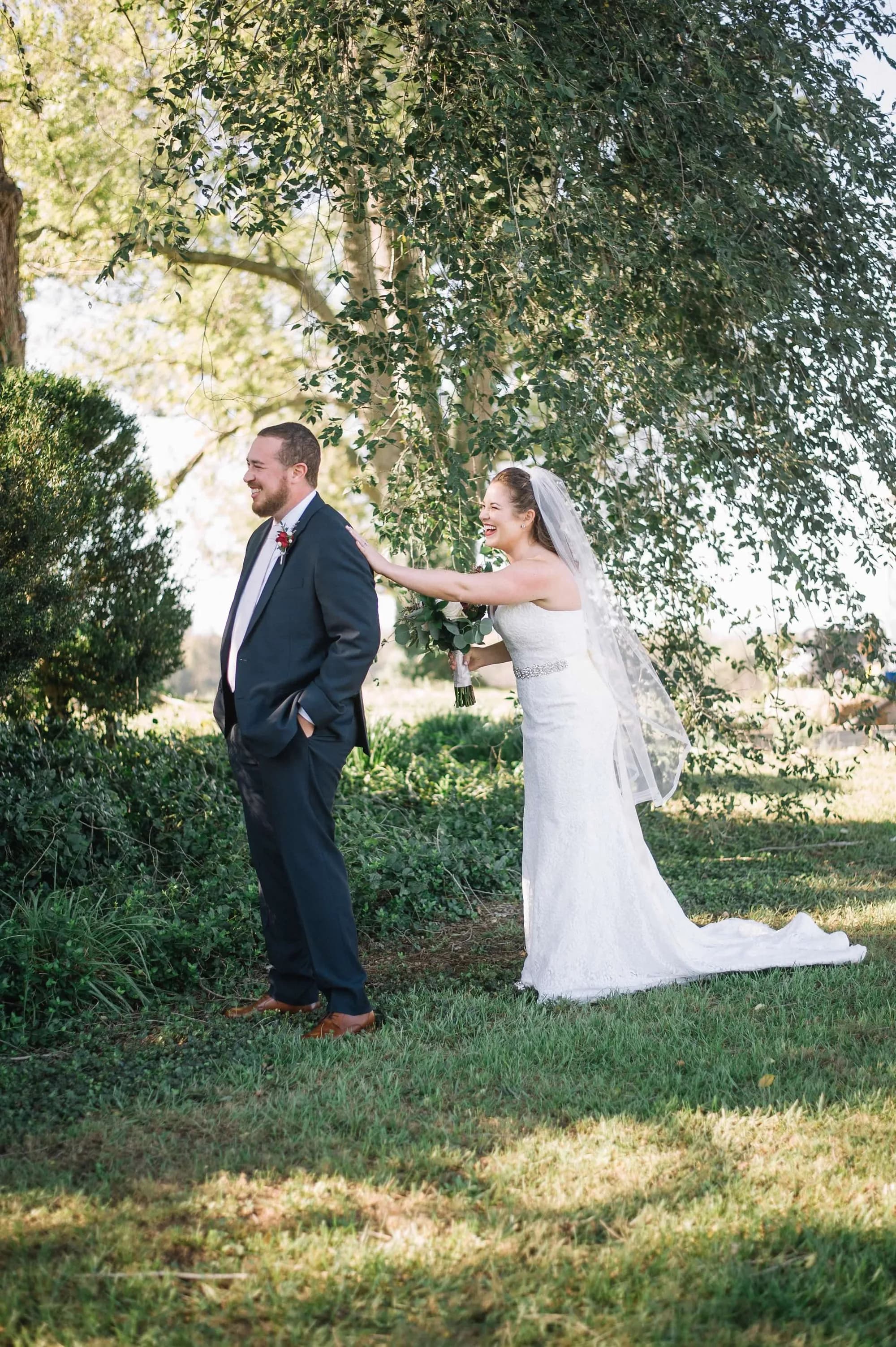 Bride laughing as she taps groom's shoulder during first look on sunny tree-lined grounds