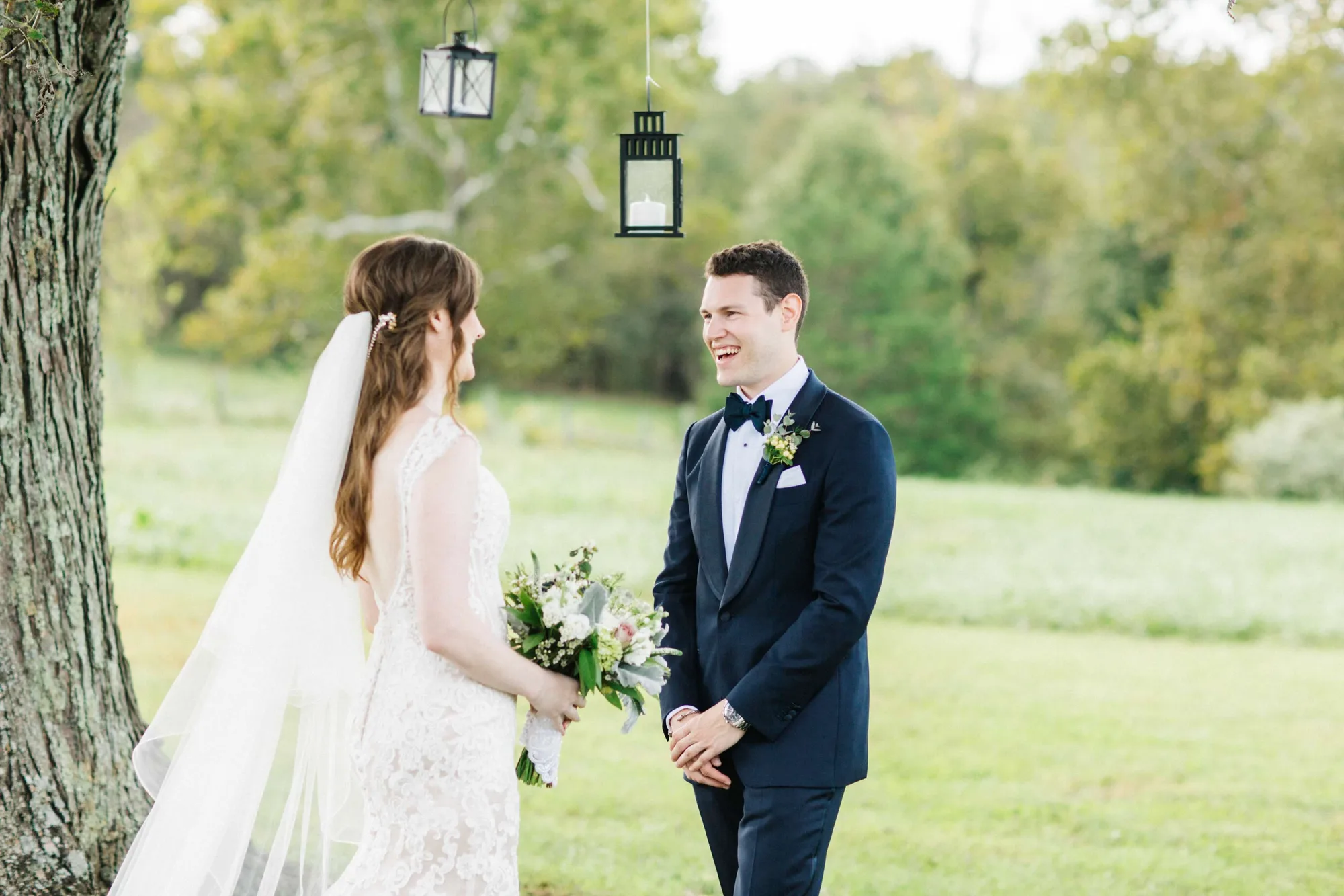 Groom laughs joyfully during first look with bride under hanging lanterns on Rixey Manor's sunlit grounds