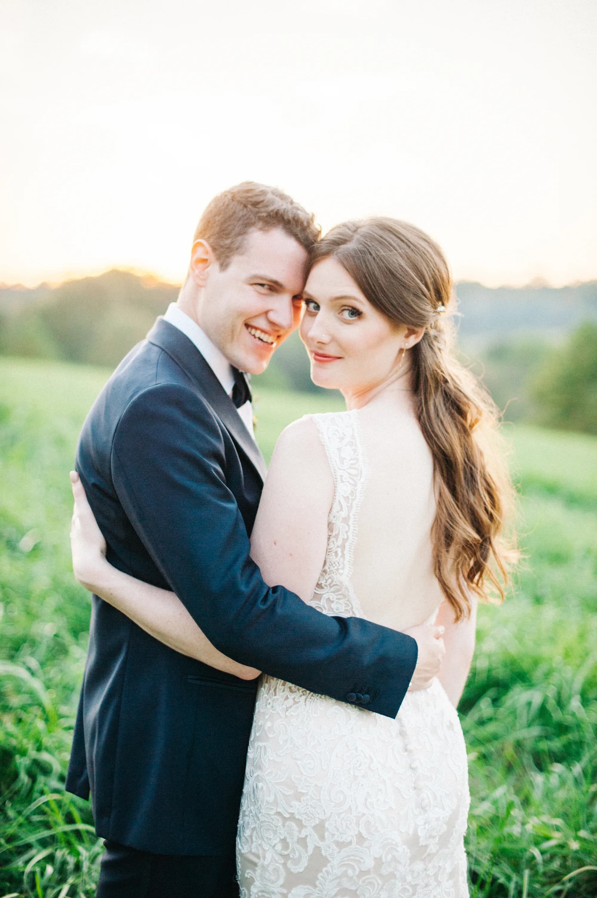 Bride and groom embrace in a grassy field at sunset, smiling at camera in romantic outdoor portrait