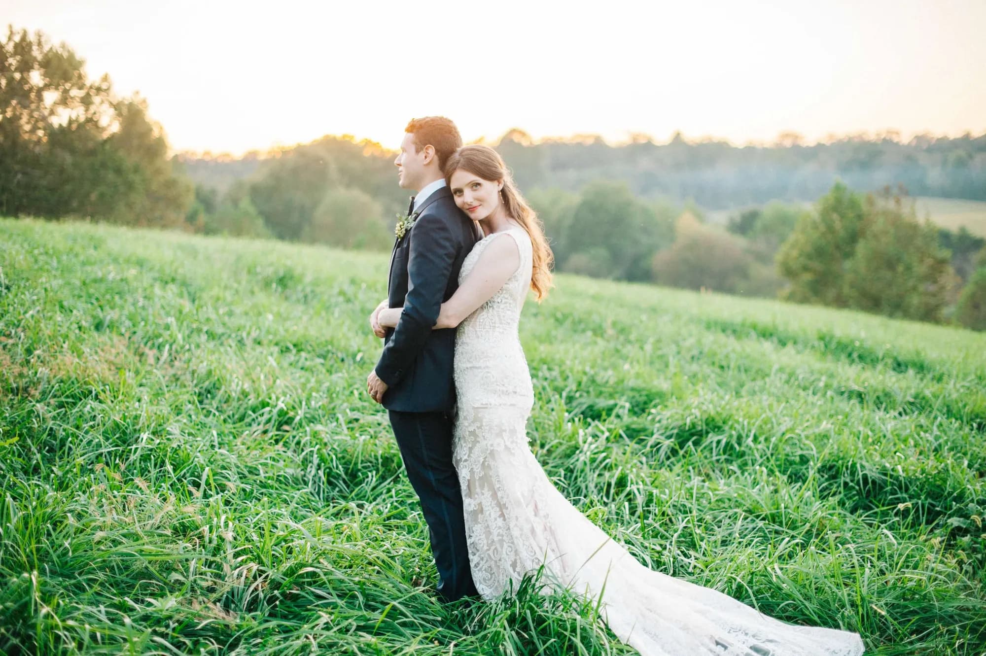 Bride embraces groom from behind on golden-hour hillside at Rixey Manor, lace gown train flowing in green fields