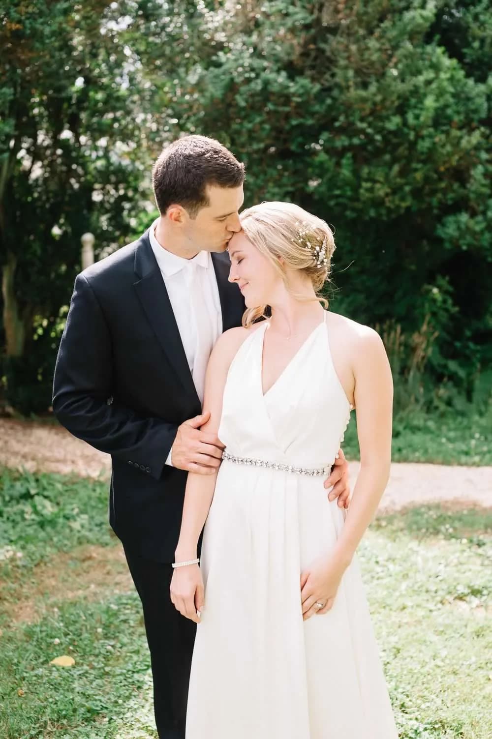Groom kisses bride's forehead as she smiles softly in a sunlit garden setting