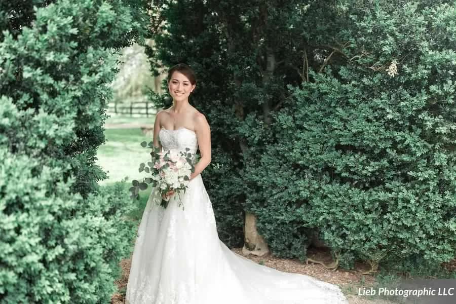 Smiling bride in lace gown holding bouquet framed by lush green hedges on Rixey Manor grounds