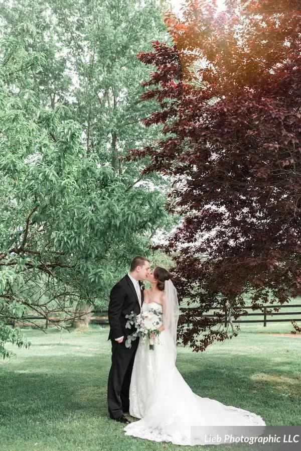 Bride and groom share a kiss beneath lush green and burgundy trees on the Rixey Manor grounds
