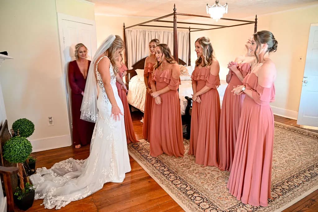 Bride reveals wedding dress to delighted bridesmaids in dusty rose gowns inside a warmly lit bridal suite