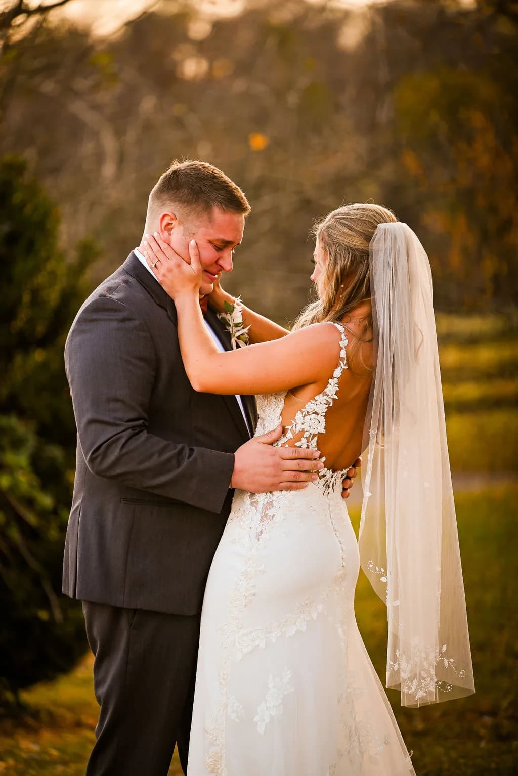 Groom tearfully embraces bride in lace gown and veil during golden hour portraits on estate grounds