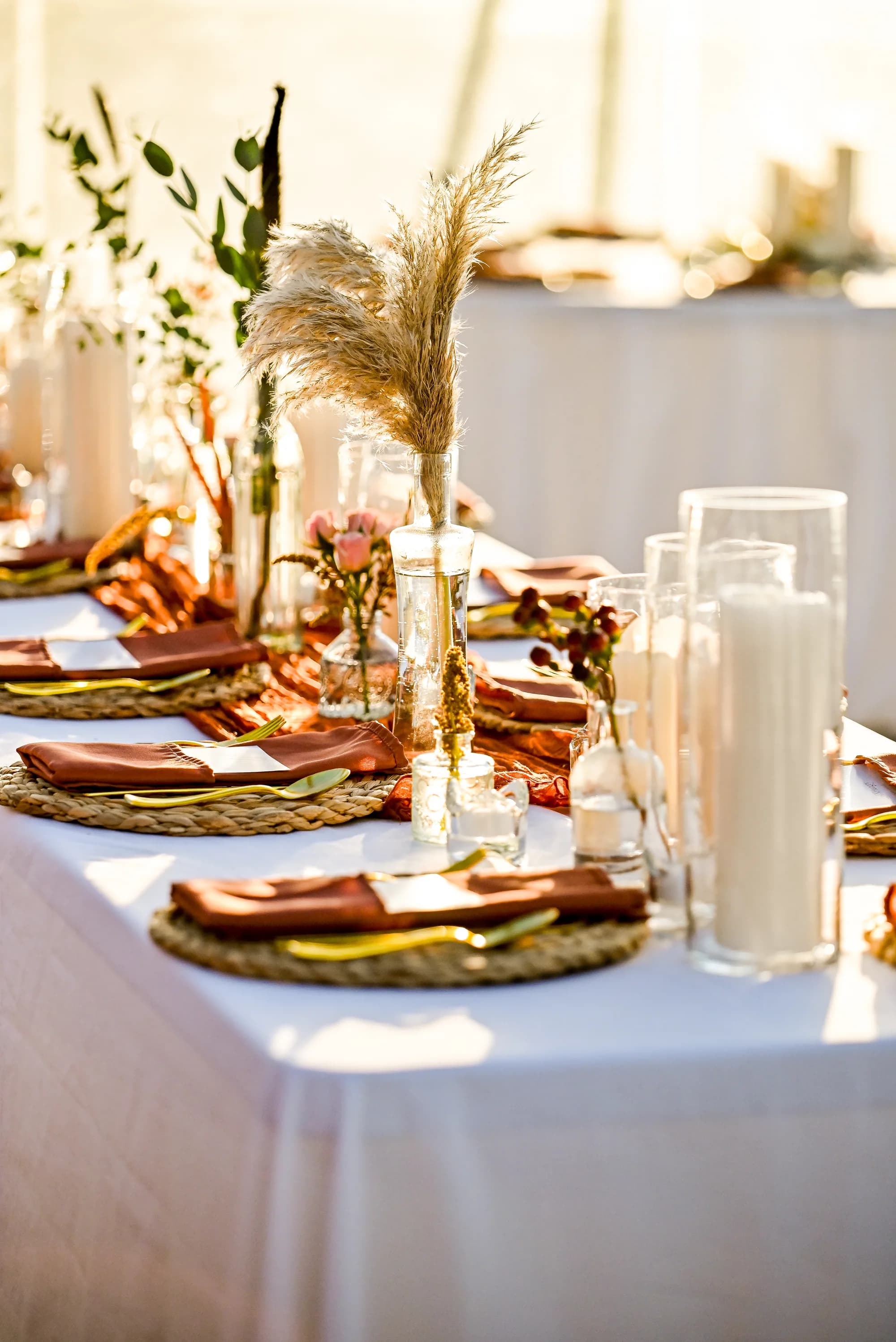 Elegant wedding reception table with white linens, gold pampas grass centerpieces, brown napkins, and white candles.
