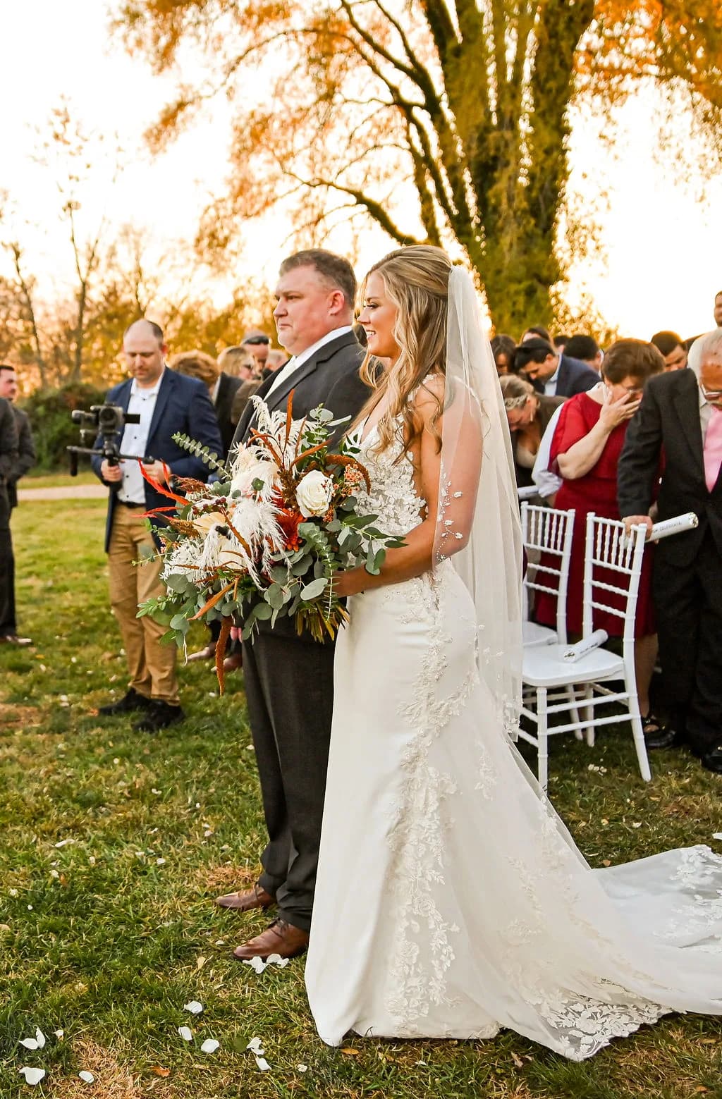 Bride in lace gown holds wild bouquet walking aisle at outdoor fall ceremony at Rixey Manor