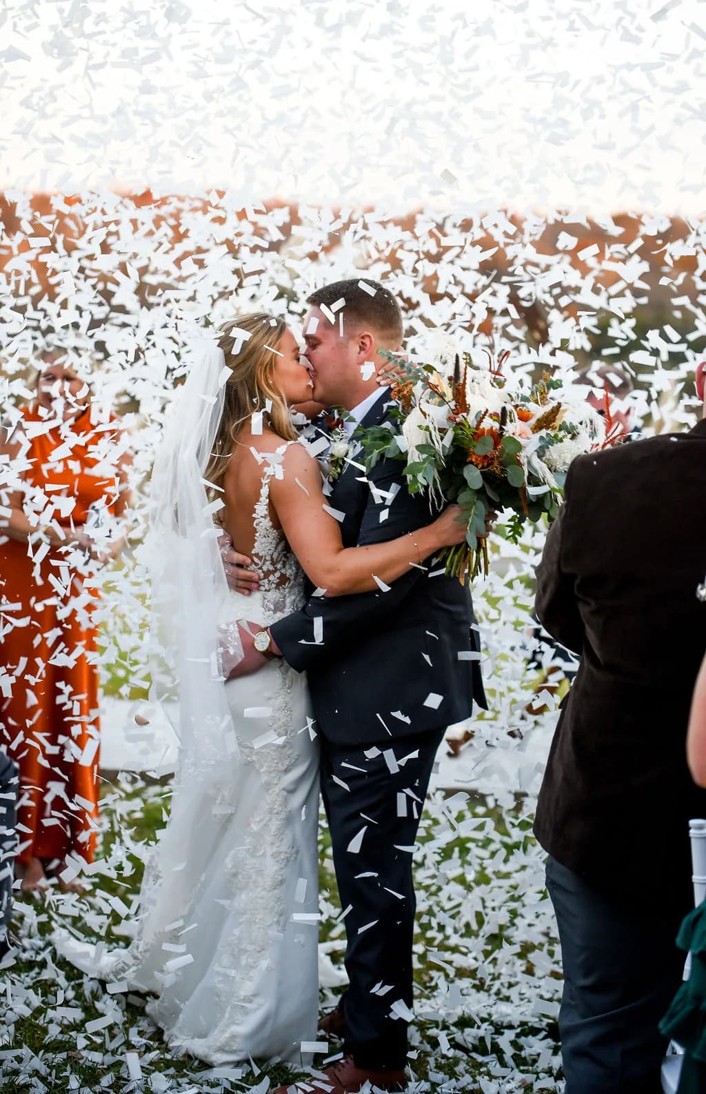 Bride and groom share first kiss surrounded by cascading white confetti at outdoor wedding ceremony