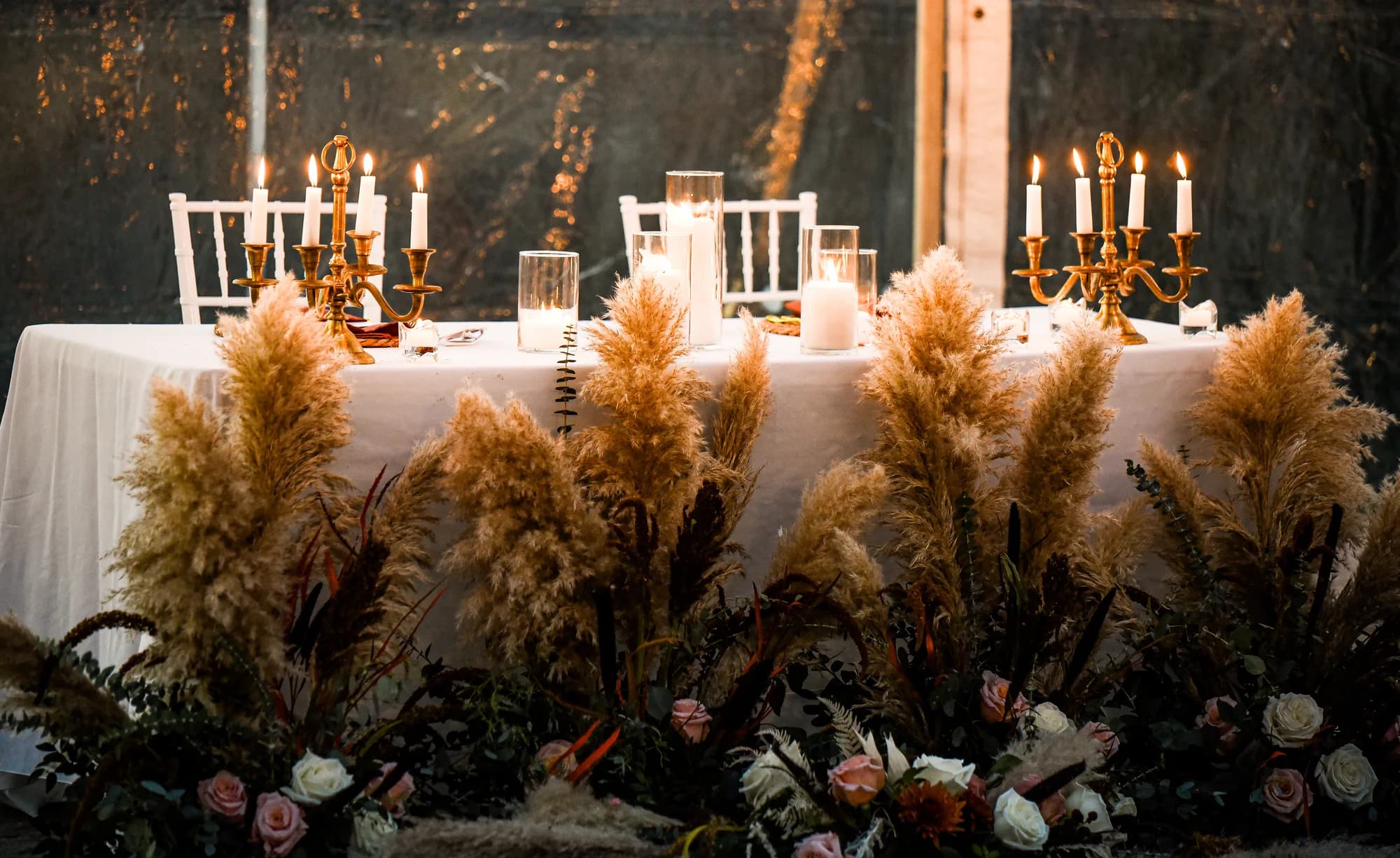 Elegant sweetheart table adorned with pampas grass, roses, and gold candelabras glowing at a wedding reception