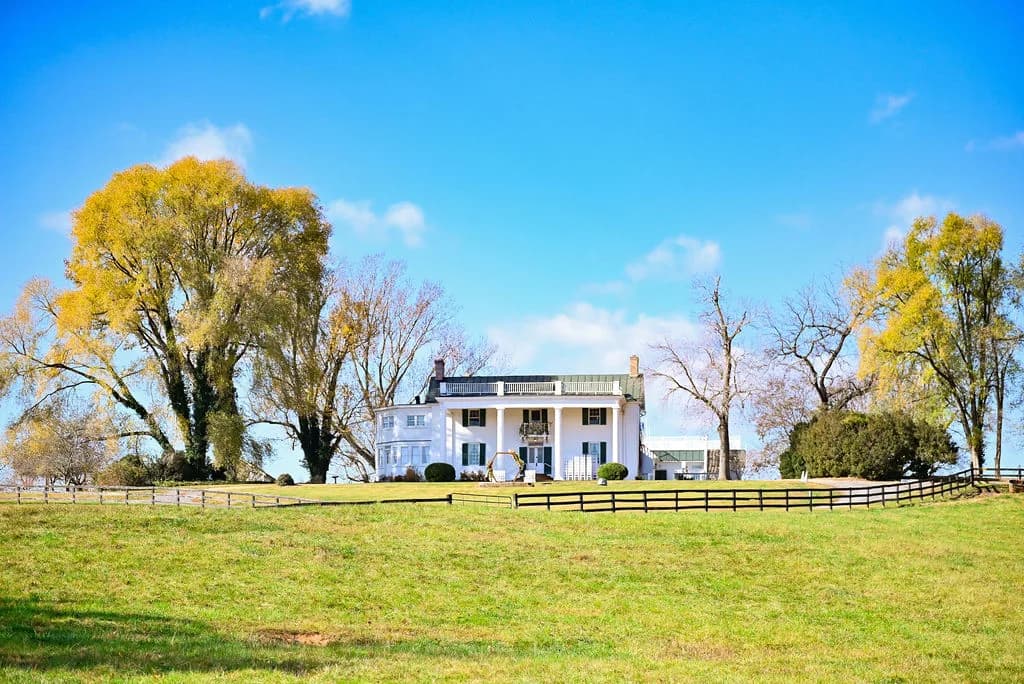 Rixey Manor historic white estate sits on a sunlit hill surrounded by autumn trees and split-rail fencing in rural Virginia