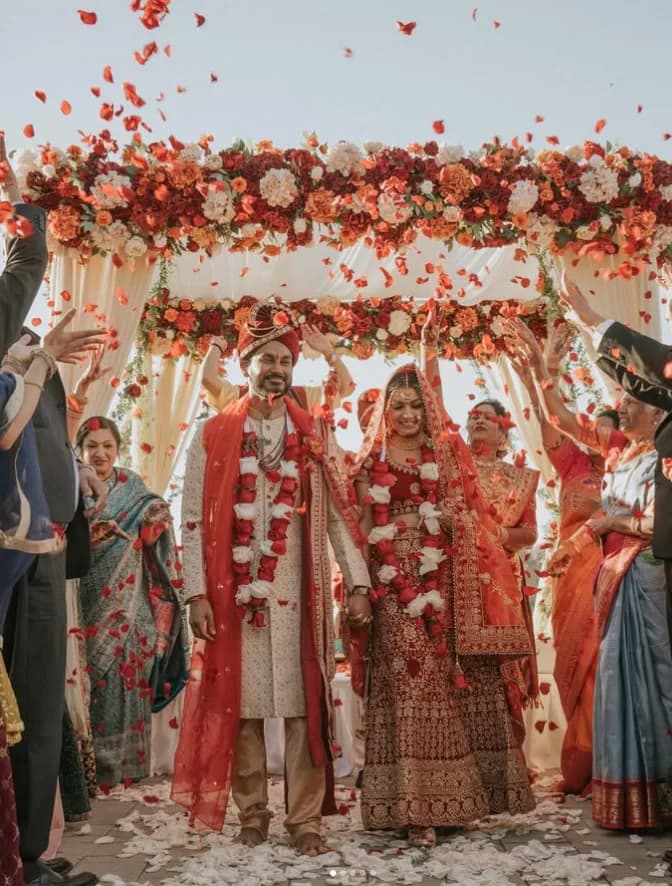 Indian bride and groom walk under floral mandap as guests shower them with red rose petals