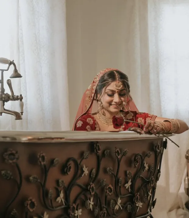 South Asian bride in red lehenga holds a rose in an ornate copper bathtub, smiling softly