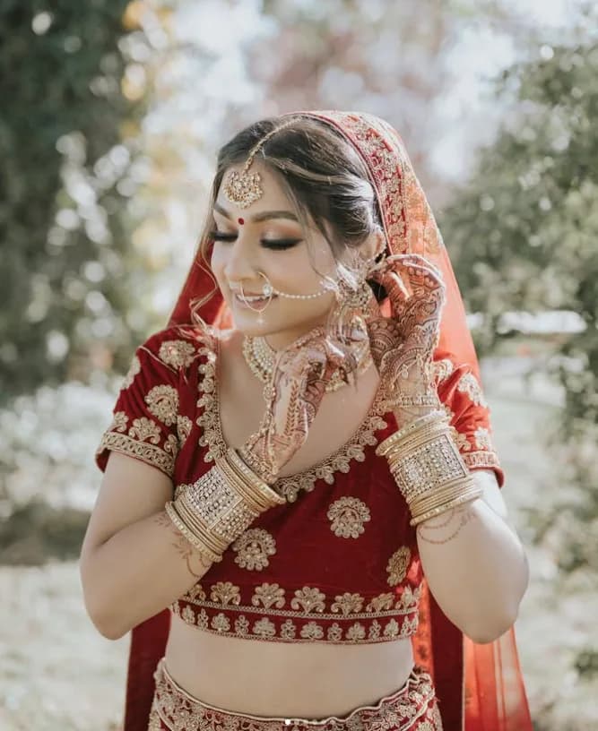 Indian bride in red and gold lehenga with ornate jewelry and mehndi, gazing downward in soft outdoor light