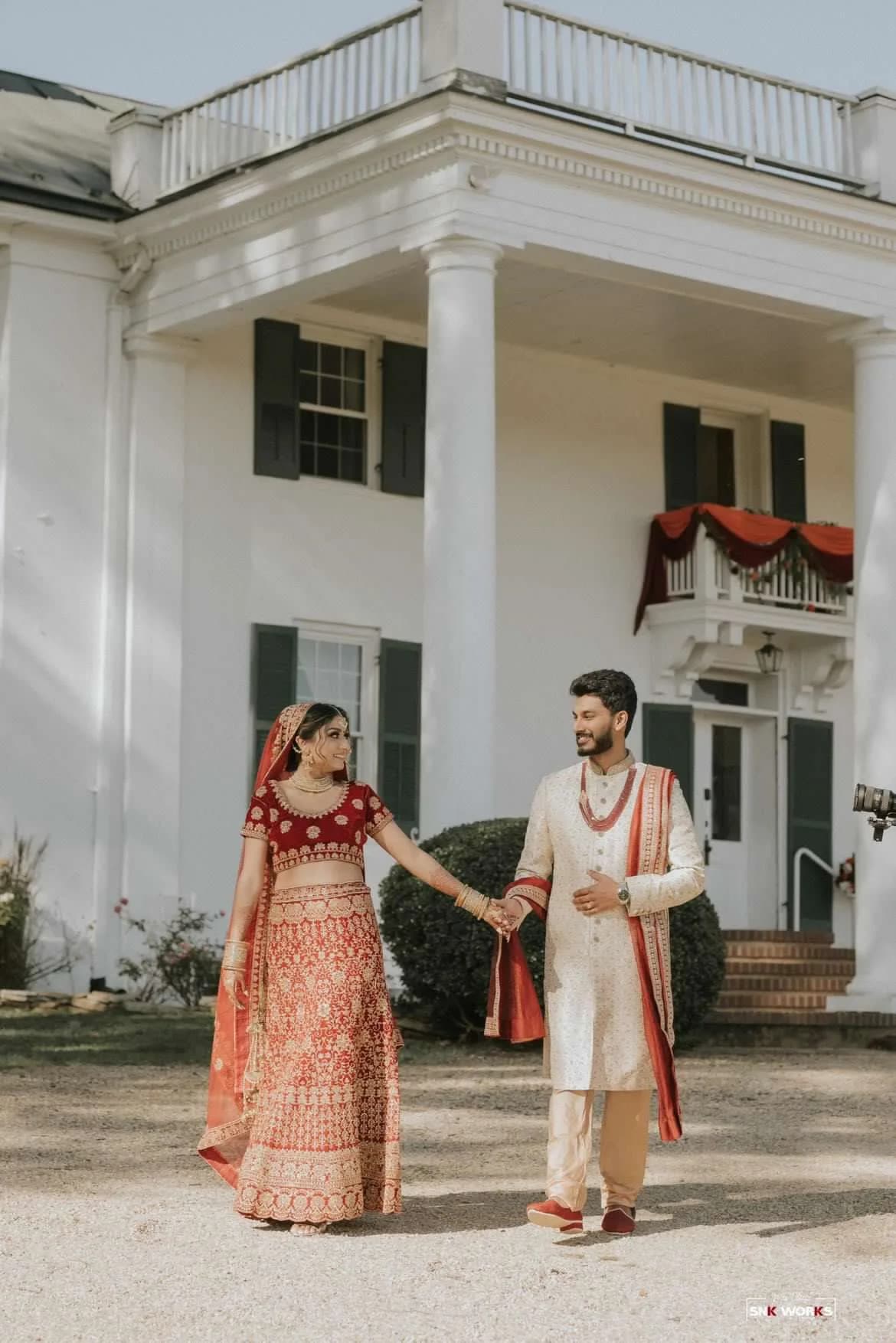 Indian bride and groom in red and gold attire hold hands in front of Rixey Manor's white columned facade
