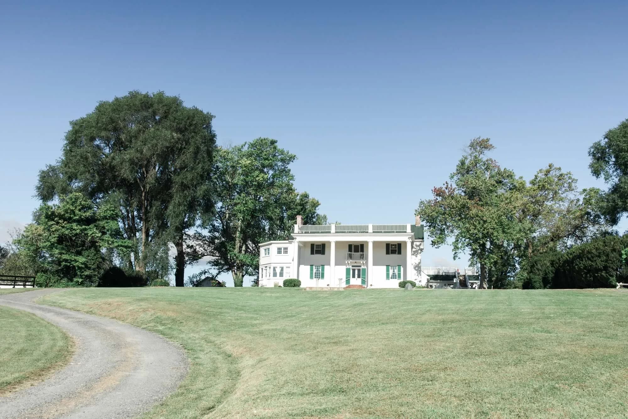 Wide view of Rixey Manor's white columned estate surrounded by mature trees and manicured lawn on a clear day.