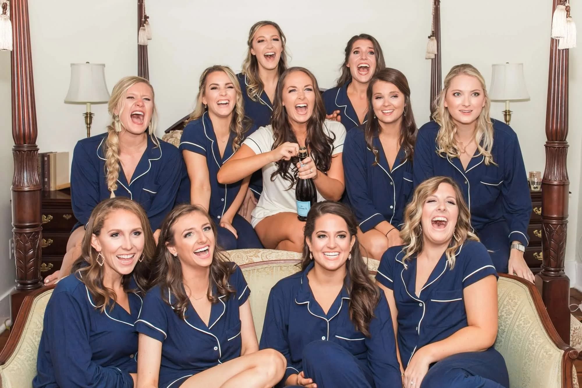 Bride in white pajamas celebrates with bridesmaids in navy sets, laughing and popping champagne in a classic bedroom