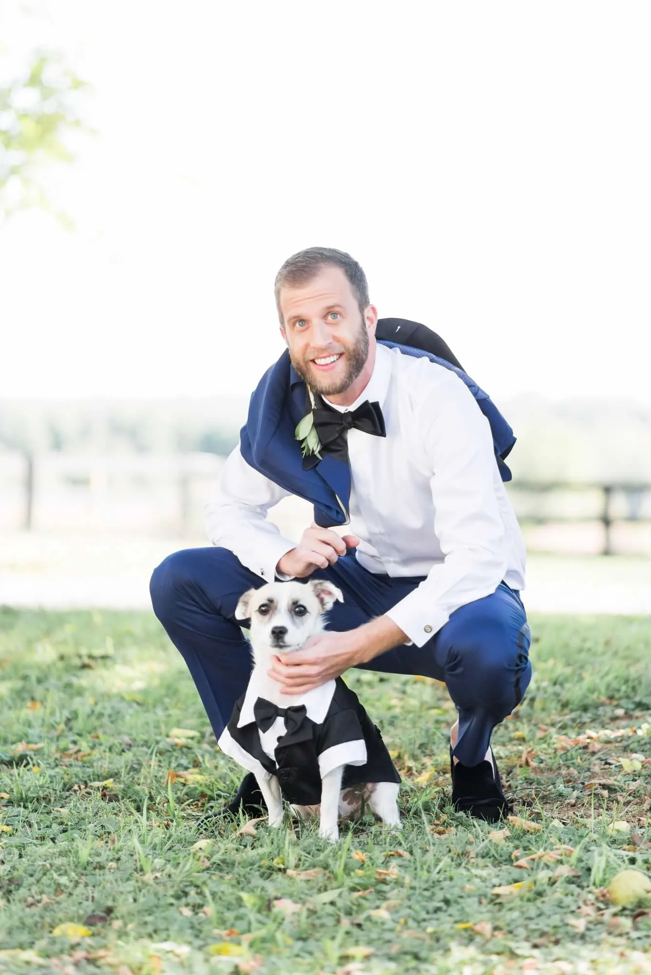 Groom in navy suit and bow tie posing with small dog in tuxedo on grass lawn