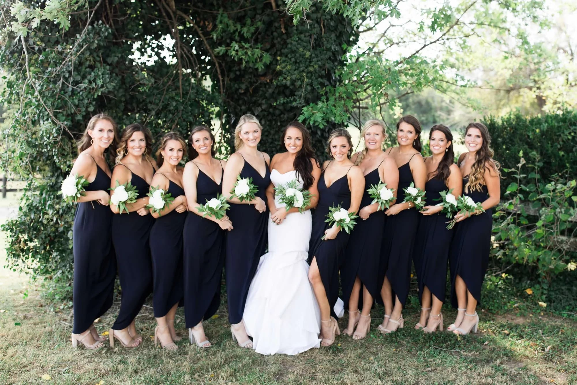 Bride in white gown surrounded by bridesmaids in navy dresses holding white bouquets outdoors