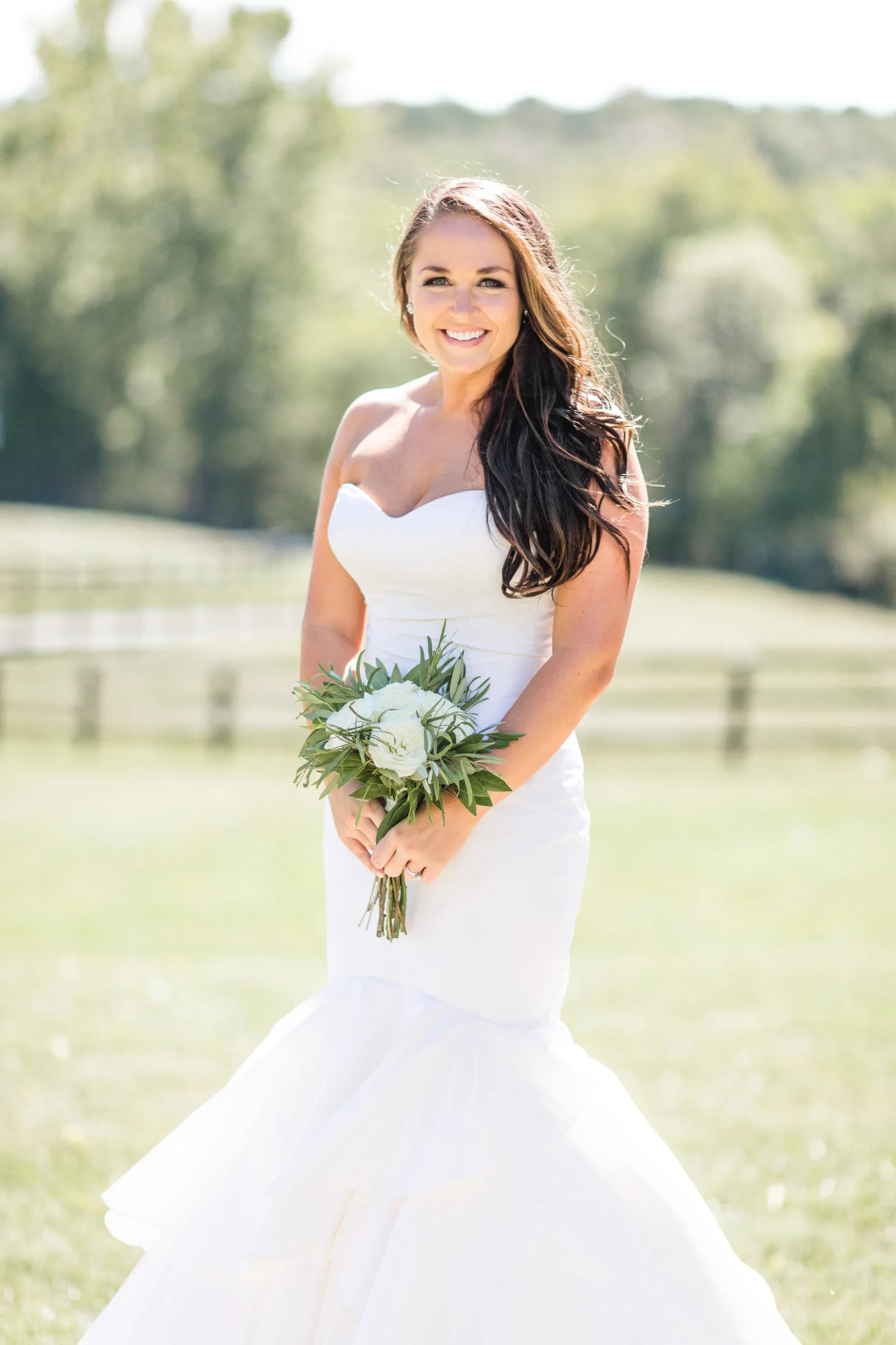 Radiant bride in mermaid gown holding greenery bouquet on sunny Rixey Manor estate grounds with pastoral fence backdrop