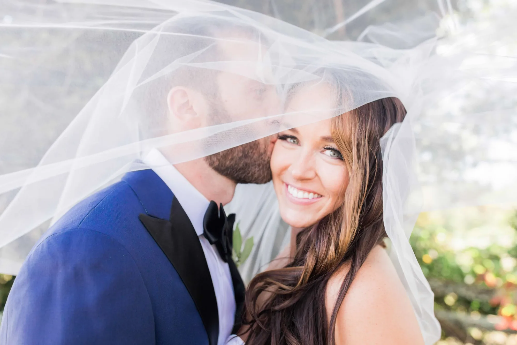 Bride smiles radiantly as groom kisses her cheek beneath flowing veil in dreamy outdoor portrait