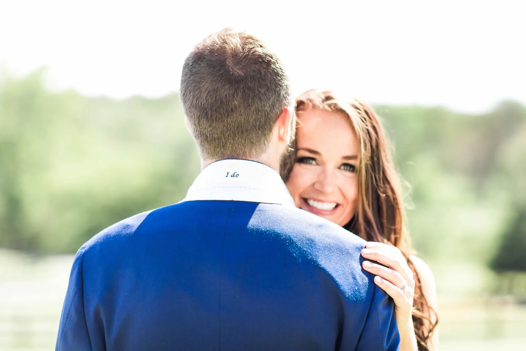 Bride smiles over groom's shoulder during outdoor couple portrait, 'I do' embroidered on his collar