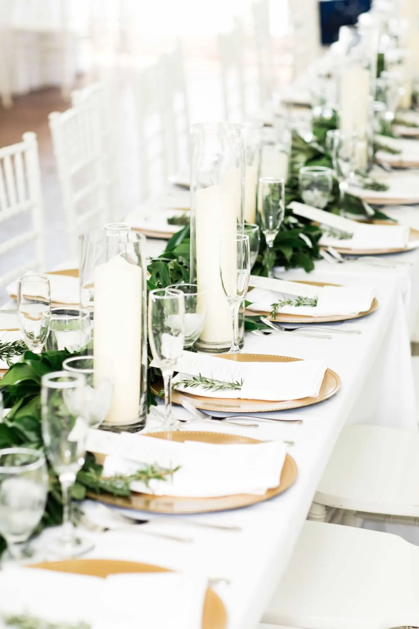 Elegant wedding reception tablescape with white pillar candles, gold chargers, and lush greenery garland runner