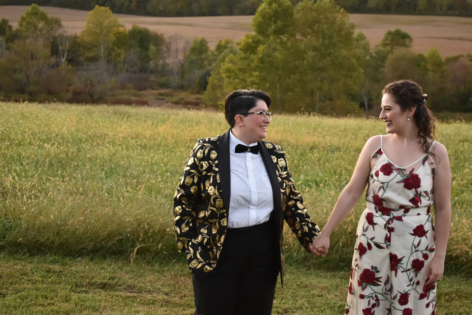 Two brides walk hand-in-hand through a golden field, smiling, one in a floral blazer and one in a rose sundress.