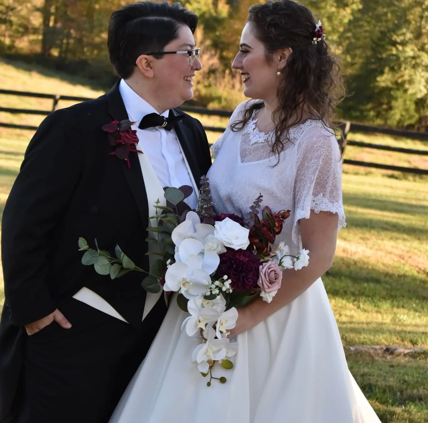 Bride and groom smiling at each other outdoors with bouquet, wooden fence and trees in background