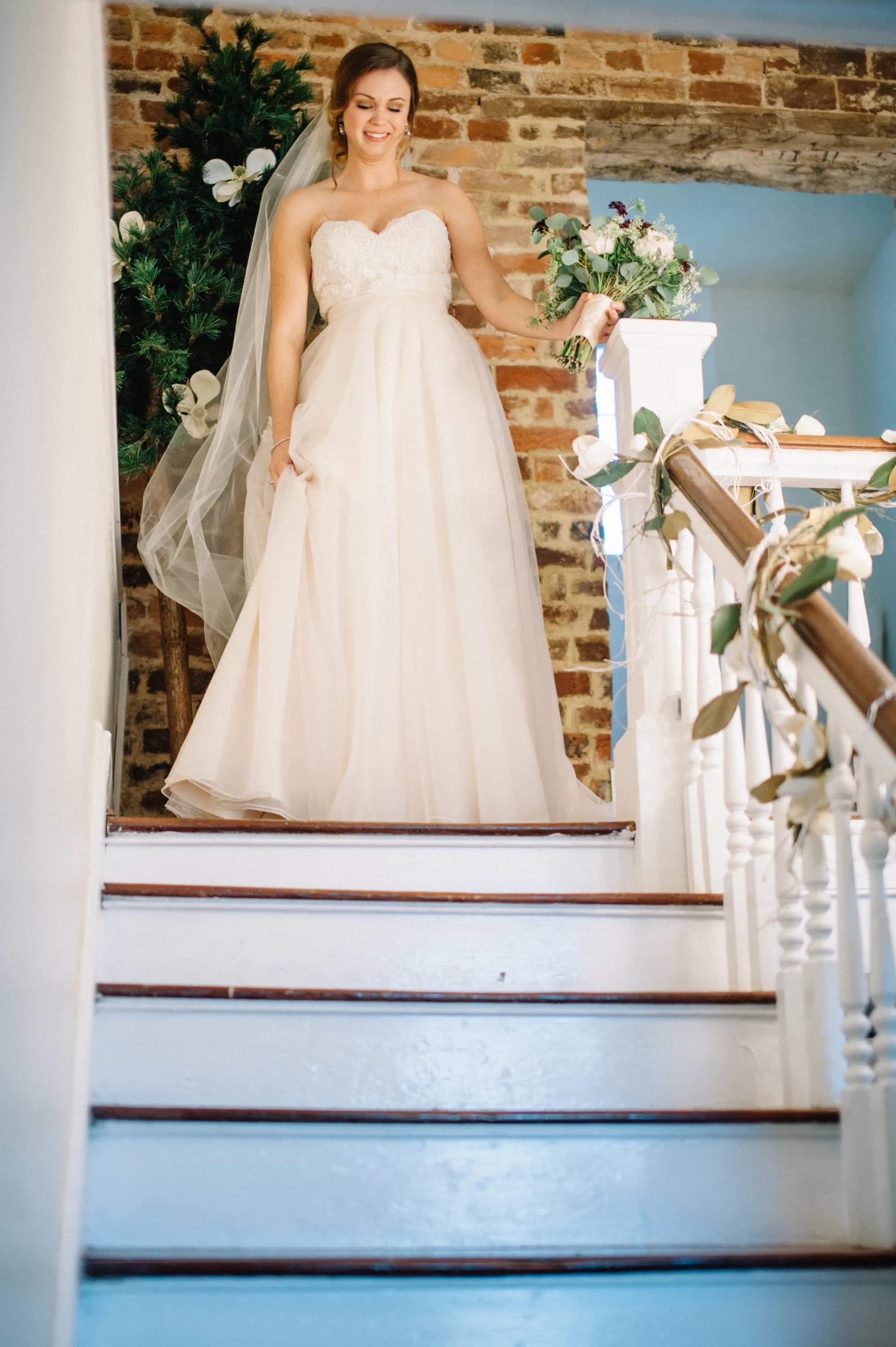 Bride descending decorated staircase at Rixey Manor, holding bouquet, smiling softly in strapless gown and veil