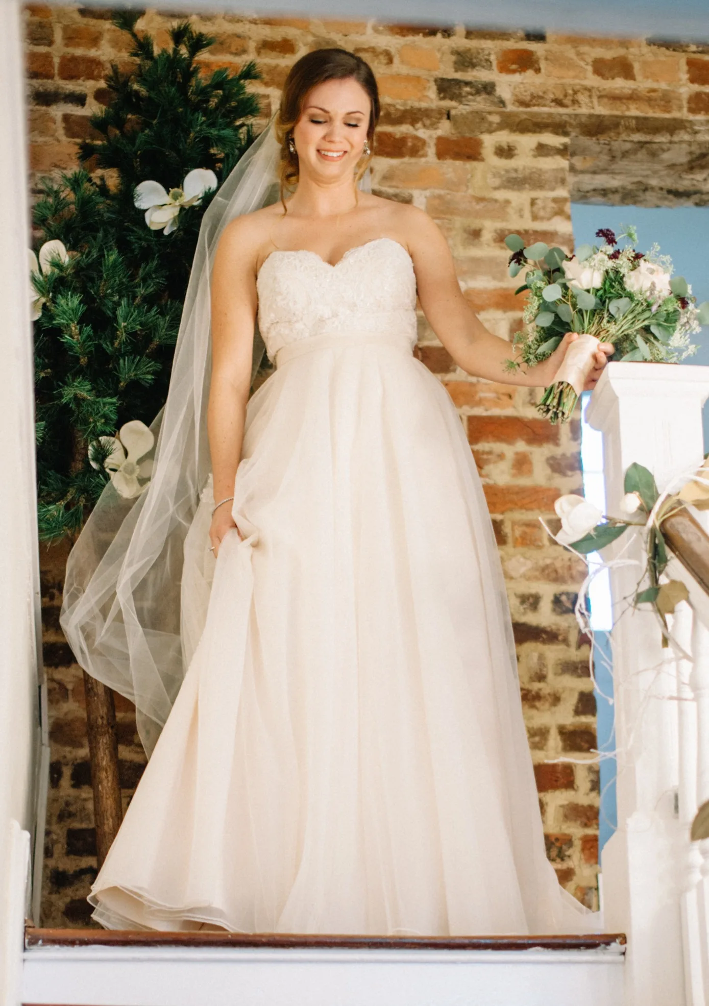 Smiling bride in strapless gown and veil descends staircase at Rixey Manor holding floral bouquet