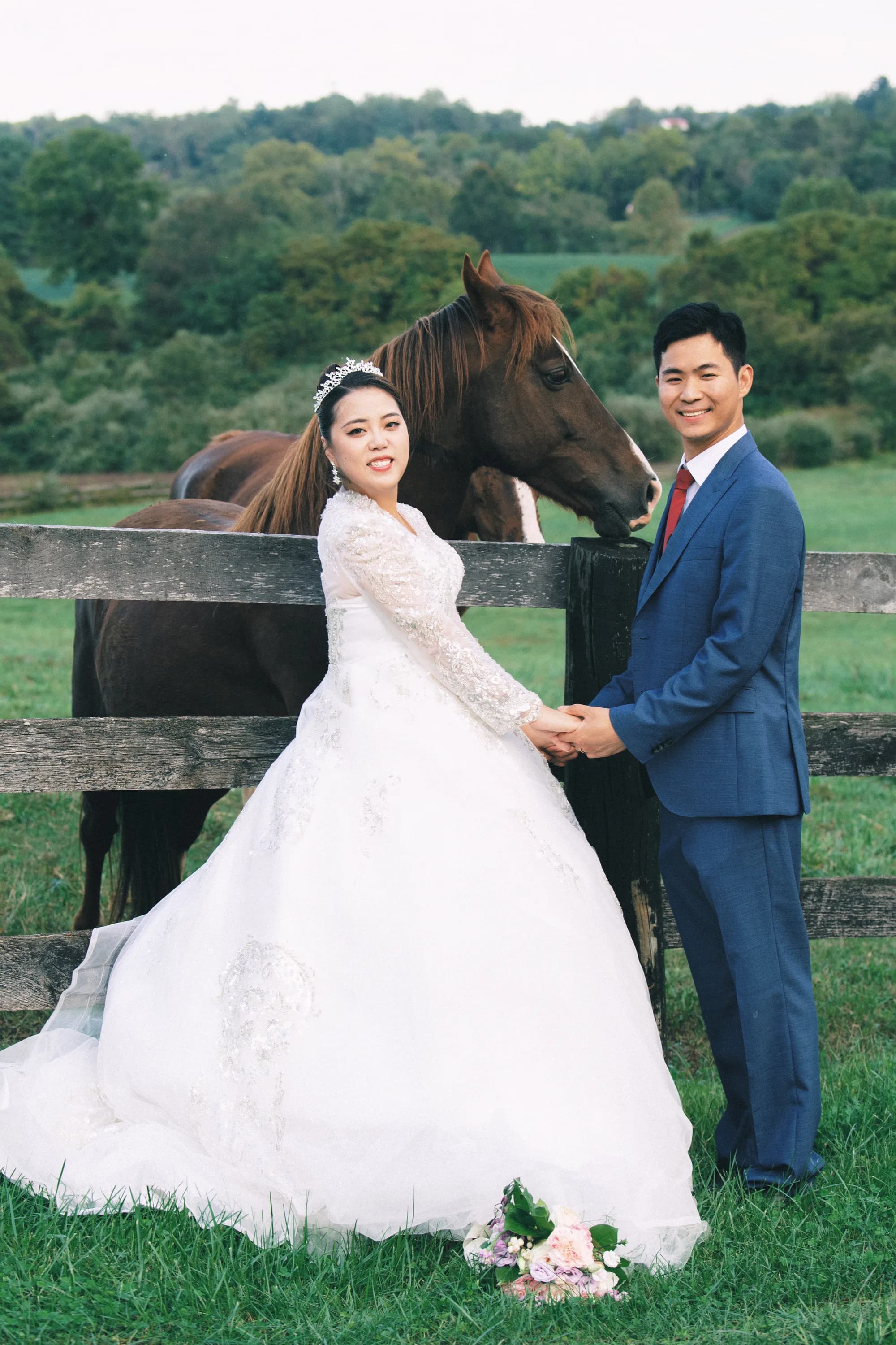 Bride and groom hold hands by a wooden fence with a chestnut horse on lush green Virginia countryside grounds