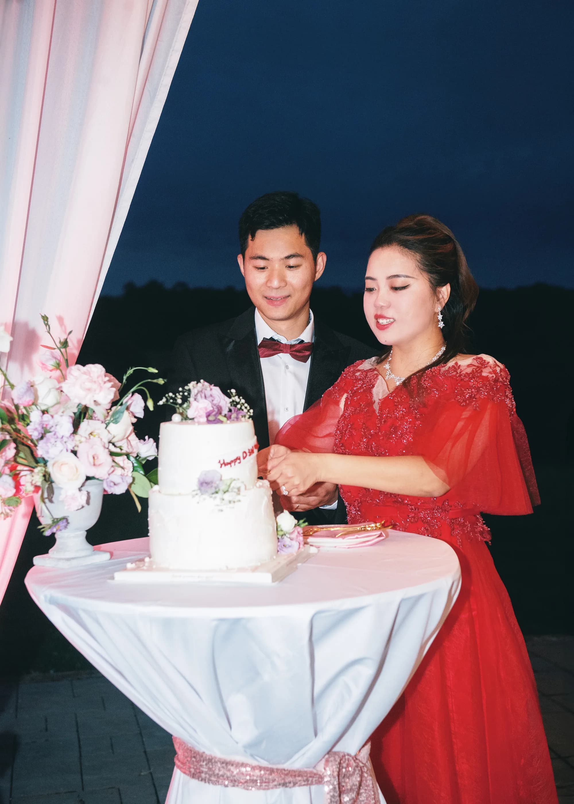 Bride in red gown and groom in tuxedo cut their two-tier wedding cake at an outdoor evening reception