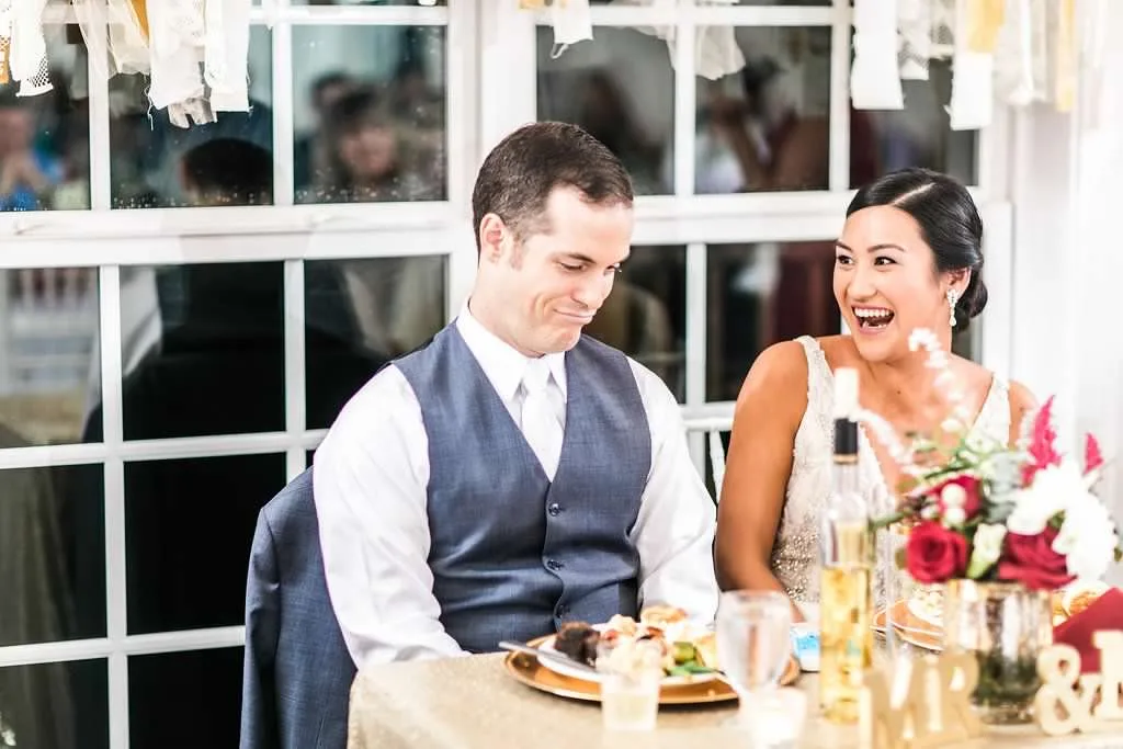 Bride laughing joyfully beside groom at sweetheart table during wedding reception toasts