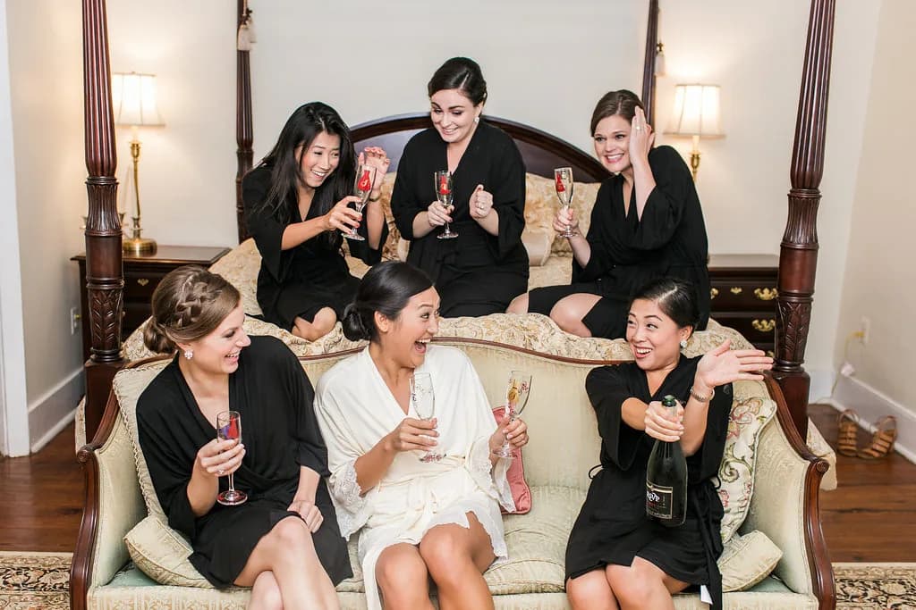 Bride in white robe laughs with bridesmaids in black robes toasting champagne in a elegant bridal suite