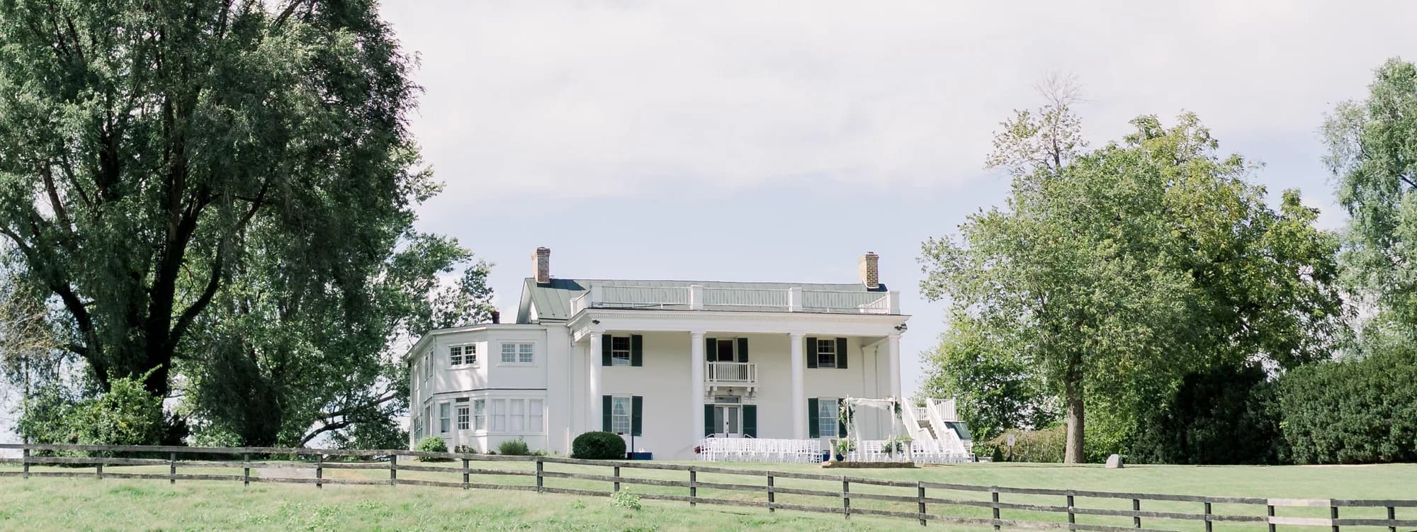 Wide exterior view of Rixey Manor's white colonial estate framed by trees and wooden fence in rural Virginia