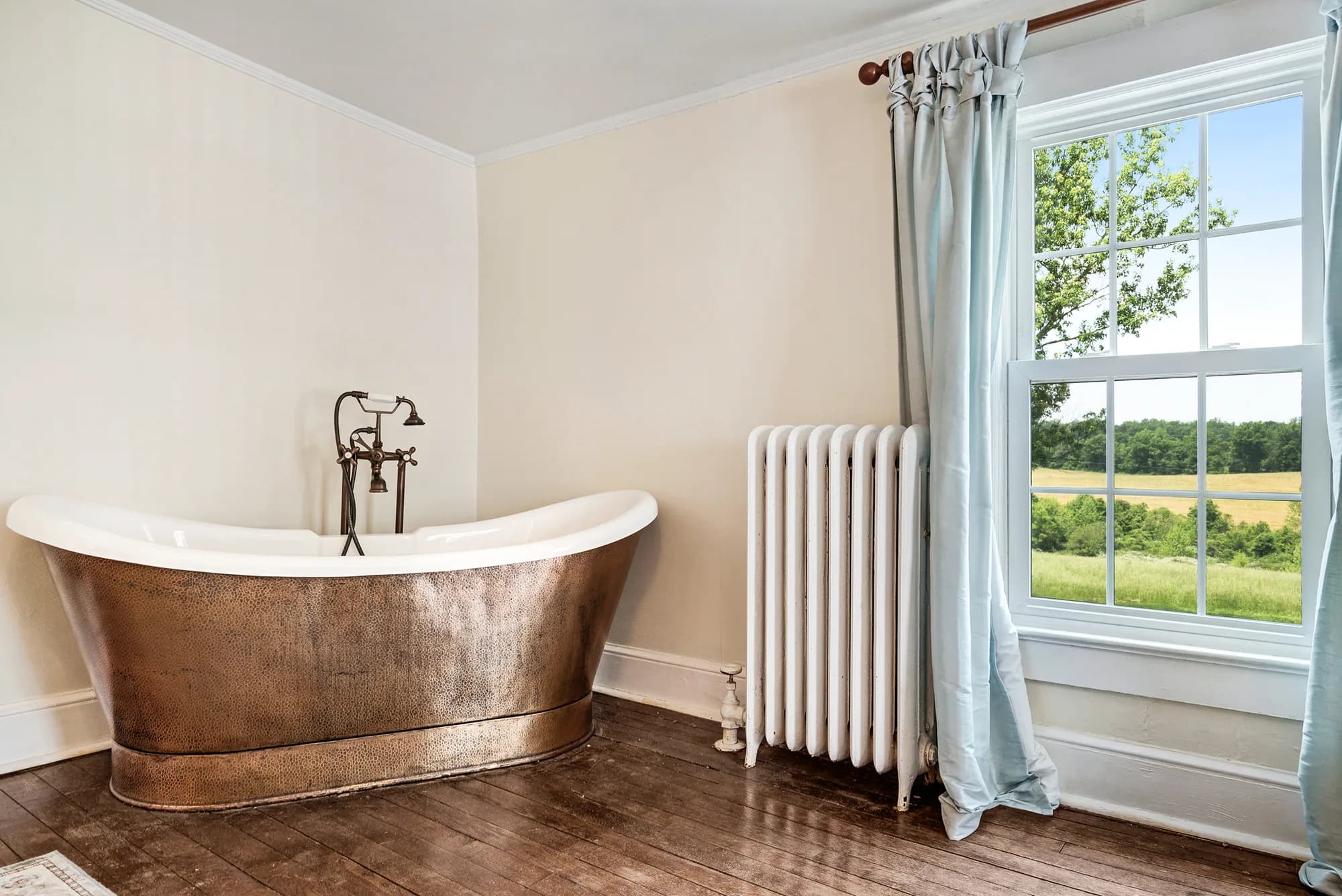 Copper clawfoot soaking tub beside a white radiator and pastoral window view at Rixey Manor