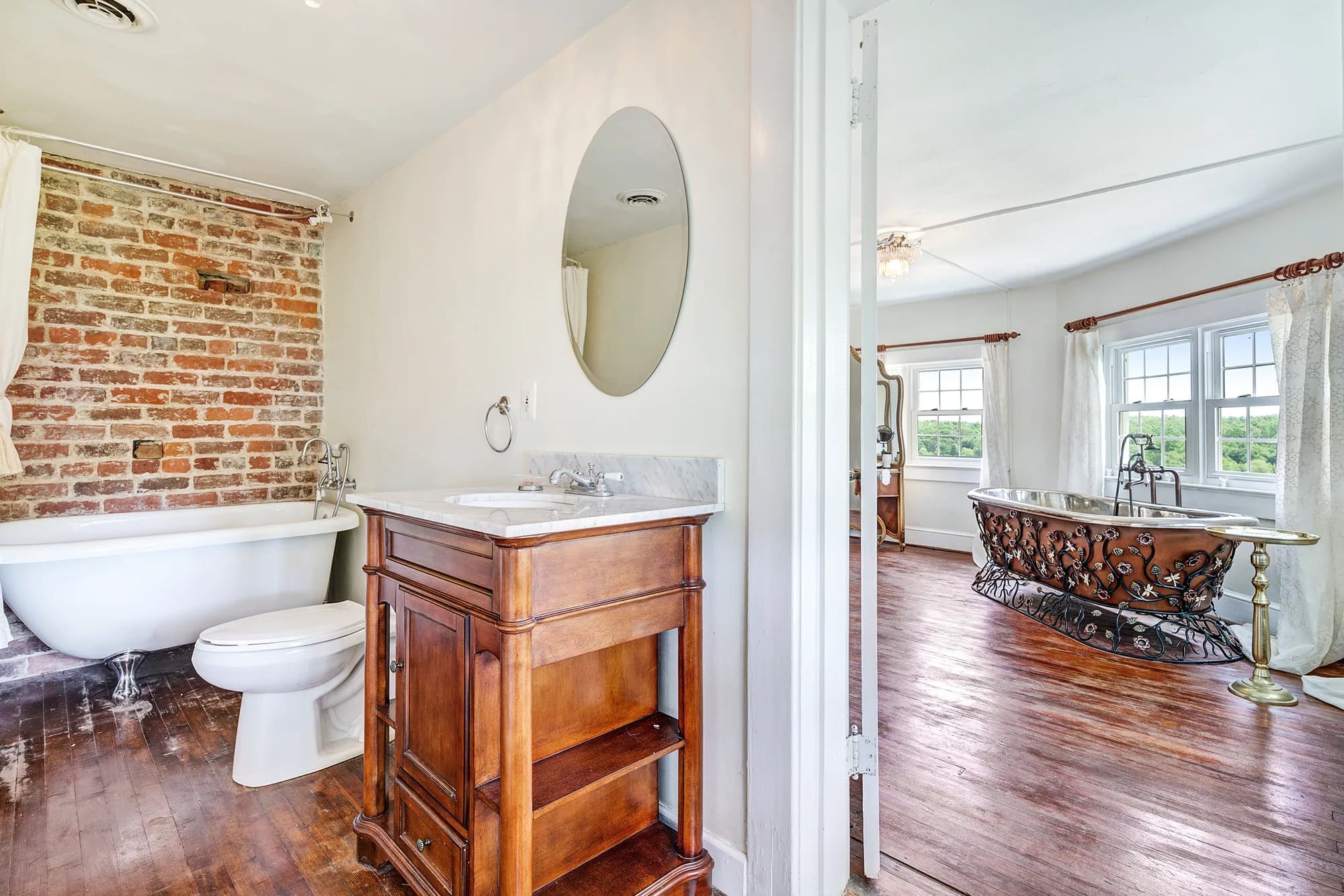 Elegant bathroom with exposed brick wall, wooden vanity, oval mirror, and view into sitting area with hardwood floors