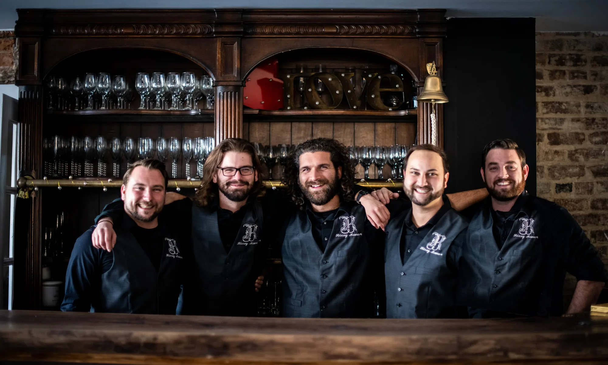 Five men in matching dark shirts posing together at a bar with wine glasses and holiday decorations